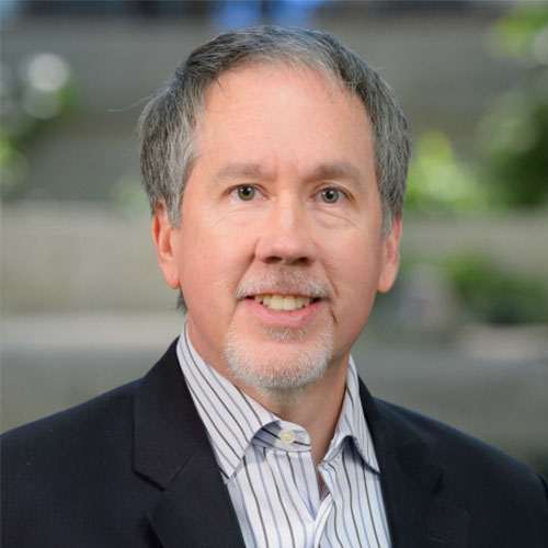 Headshot of Argonne researcher David Martin. He smiles and wears a white button up with a gray and blue line pattern. He wears a black blazer. He stands in front of a hazy nature backdrop.