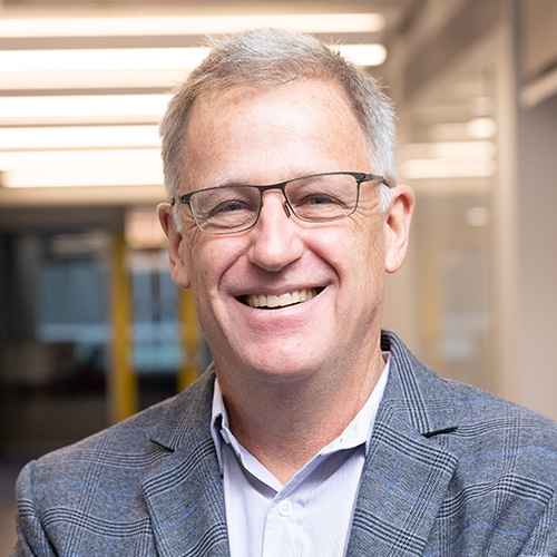 Headshot of Argonne researcher Ian Foster. He smiles and wears a black white button-up with light gray blazer. He wears black wire frame glasses. He stands in front of an office backdrop.