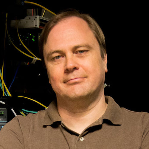 Headshot of Jefferson Laboratory researcher Ilya Baldin. He smiles and wears a brown polo. He stands in front of a piece of computing machinery.