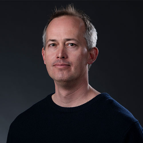 Headshot of Los Alamos researcher Earl Lawrence. He smiles and wears a dark navy sweater. He stands in front of a black backdrop.