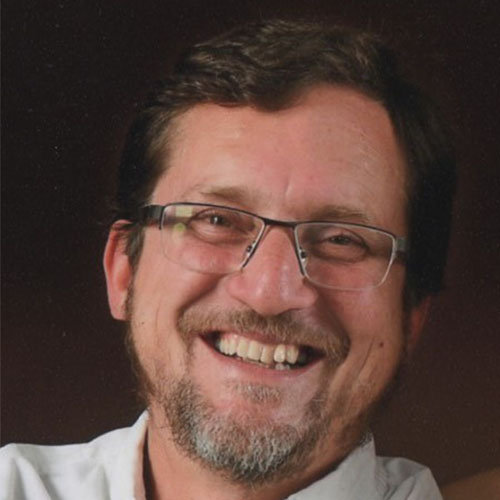 Headshot of Lawrence Berkeley researcher John Shalf. He smiles and wears a white button up. He stands in front of a black backdrop.