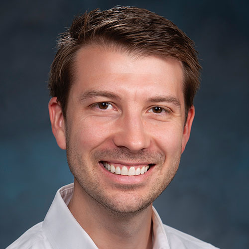 Headshot of Lawrence Livermore researcher Luc-Peterson. He smiles and wears a white button up. He stands in front of a gray backdrop.