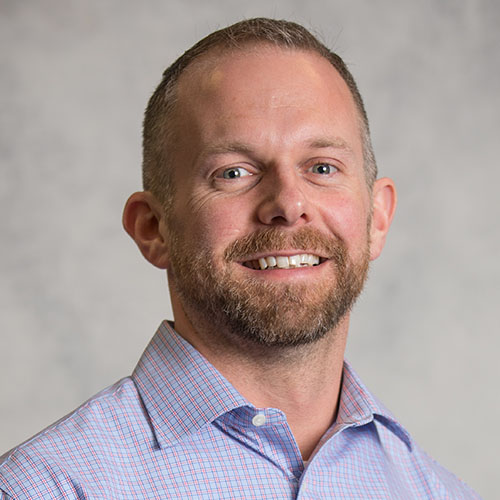 Headshot of National Energy Technology Laboratory researcher Jordan Musser. He smiles and wears a light blue button up with pink plaid squares. He stands in front of a gray backdrop.