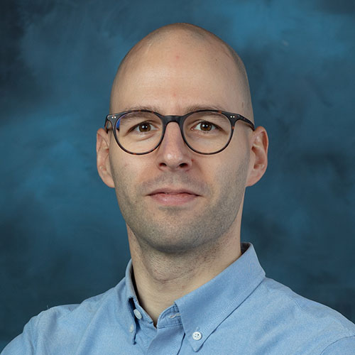 Headshot of Oak Ridge researcher Matthias Maiterth. He smiles and wears a light blue button up. He stands in front of a dark blue backdrop.