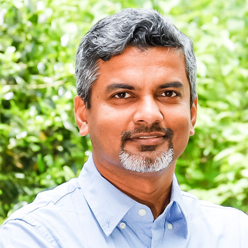 Headshot of Oak Ridge researcher Ramanan Sankaran. He smiles and wears a light blue button up. He stands in front of a hazy nature backdrop.