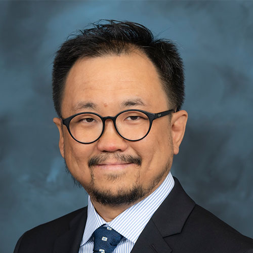 Headshot of Oak Ridge researcher Woong Shin. He smiles and wears a white button up with blue stripes. He has on a navy tie and a black blazer. He stands in front of a dark blue backdrop.