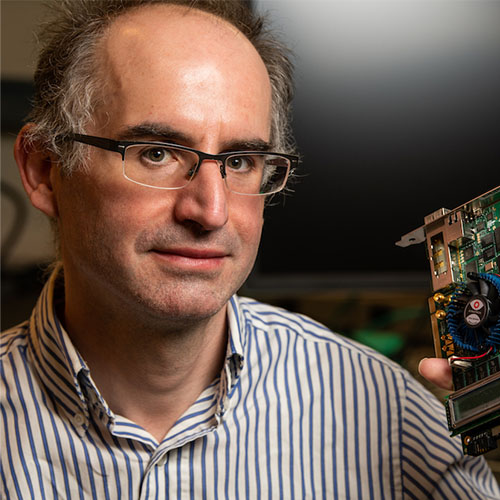 Headshot of Pacific Northwest National Laboratory researcher Antonino Tumeo. He smiles and wears a blue and white striped button-up. He holds a piece of a super computer.