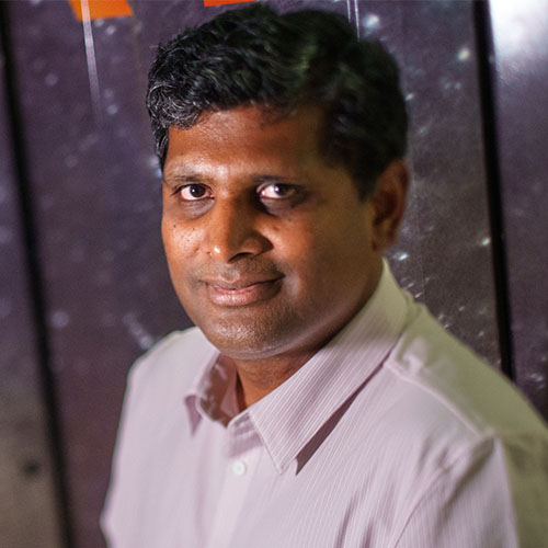 Headshot of Sandia researcher Siva Rajamanickam. He smiles and wears a light gray button up with white stripes. He stands in front of the doors of a super computer.