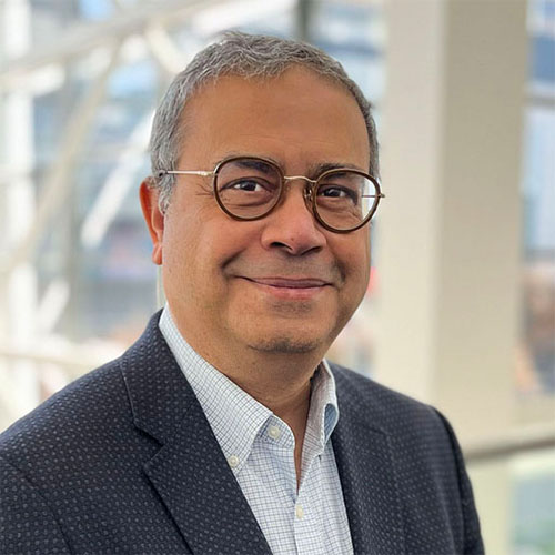 Headshot of ESnet researcher Yatish Kumar. He smiles and wears a white dress shirt with blue lines and a matching black blazer with patterning. He stands in front of a blurred interior background.