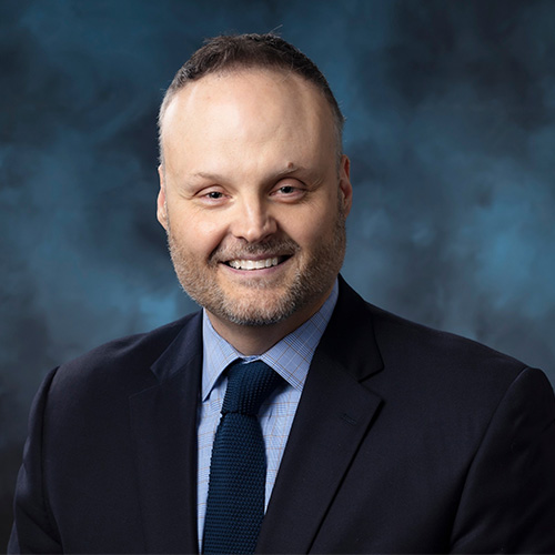 Headshot of Oak Ridge Director of Science Bronson Messer. He smiles and wears a light blue button-up with a dark navy tie and blazer. He stands in front of a teal backdrop.