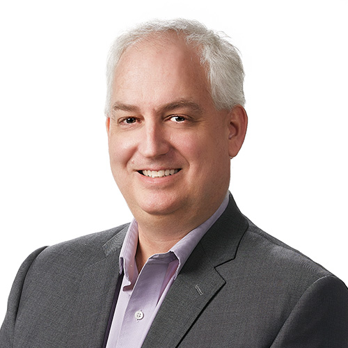 Headshot of Oak Ridge researcher Robert Neely. He smiles and wears a dark gray blazer with liac button-up. He stands in front of a white backdrop.